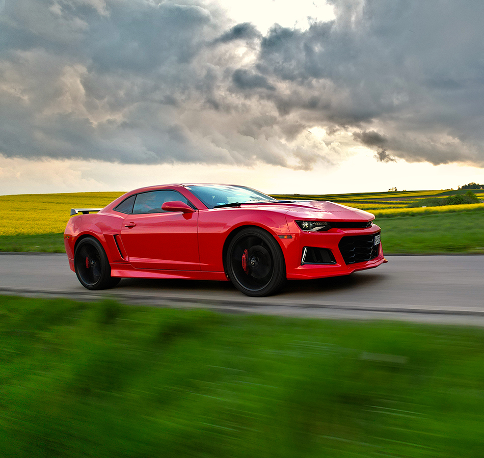 chevrolet-camaro-zdjecie-dronem Czerwony Chevrolet Camaro V8 na pierwszym planie, na tle pola rzepaku i zachodzącego słońca. Zdjęcie przy użyciu drona, Usługi dronem w branży motoryzacyjnej. profesjonalne usługi dronem i aparatem