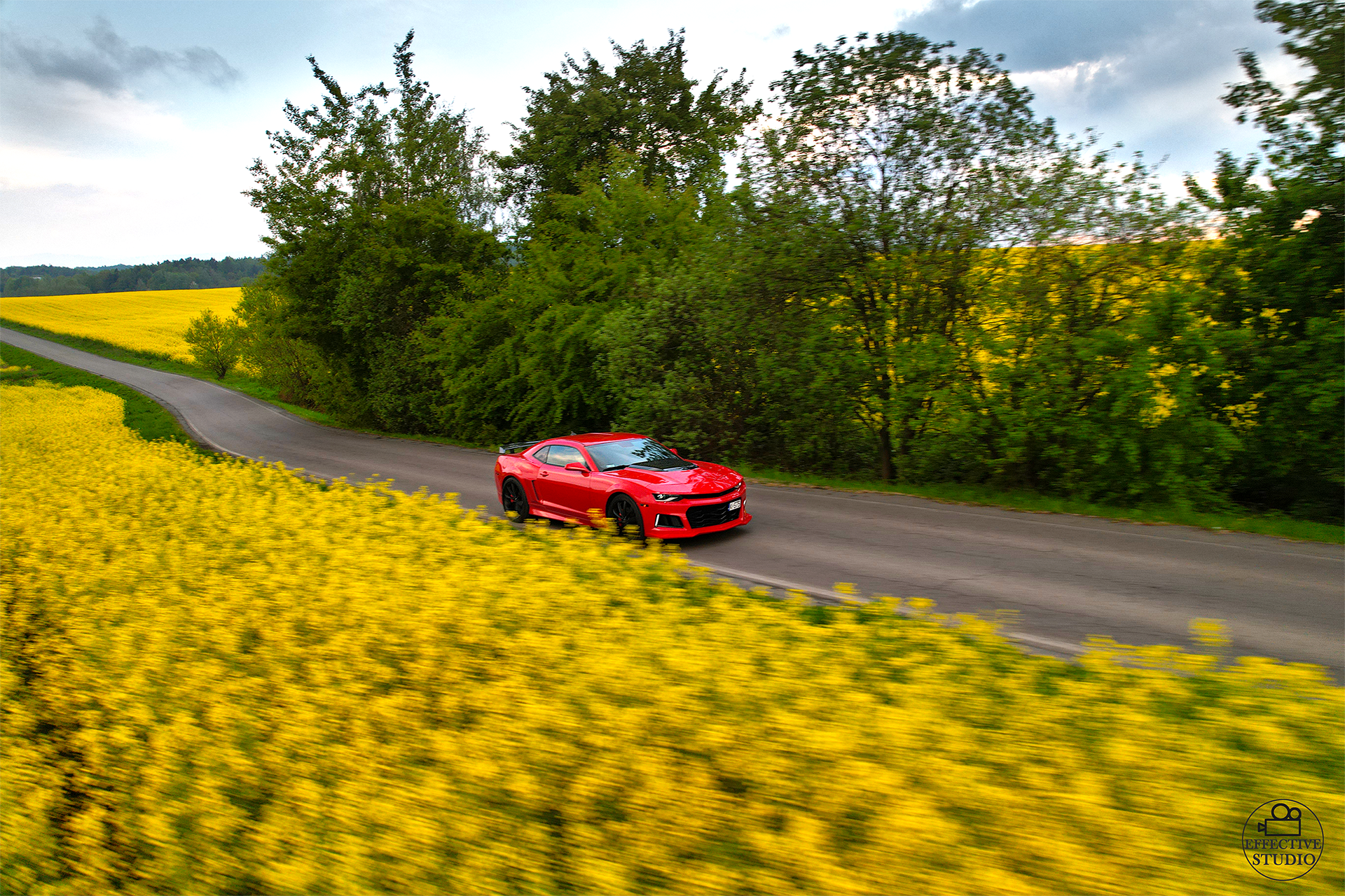 Czerwony Chevrolet Camaro z silnikiem V8 jadący drogą na tle żółtego rzepaku, zdjęcia dronem. Zdjęcia reklamowe z drona dla warsztatu samochodowego.